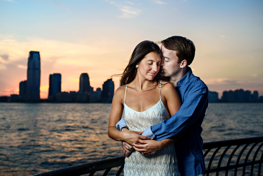 A couple embrace along the hudson river at sunset during their battery park engagement session.